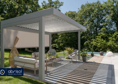 Uno de los modelos de pérgolas bioclimáticas de Abrisol. Una pérgola de color beige en un jardín decorada con muebles de exterior y en la que se puede observar que hay bastante sombra.