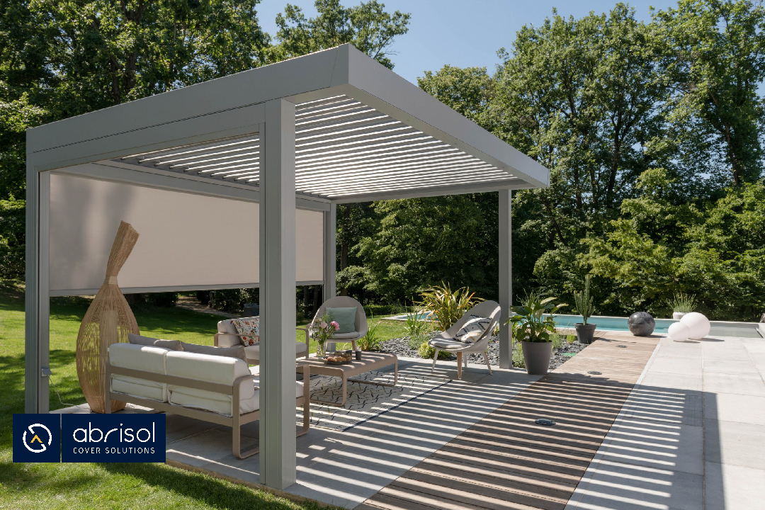 Uno de los modelos de pérgolas bioclimáticas de Abrisol. Una pérgola de color beige en un jardín decorada con muebles de exterior y en la que se puede observar que hay bastante sombra.